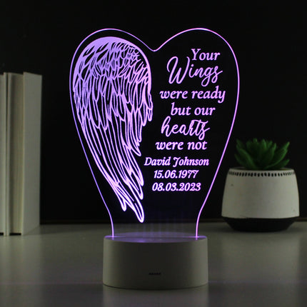 A glowing LED lamp shaped like an angel wing with a heart in the center. The light is purple, and etched into the heart are the words: "Your wings were ready but our hearts were not." Next to the lamp on the right is a small potted succulent.